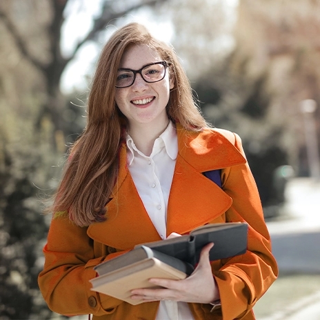 A woman with long brown hair and glasses is smiling while wearing an orange blazer.