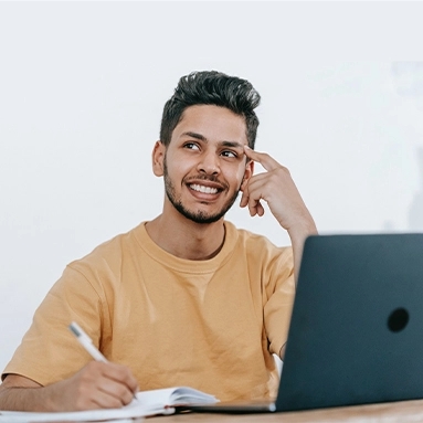 A man with short hair and a beard is smiling at a laptop while wearing a casual shirt.